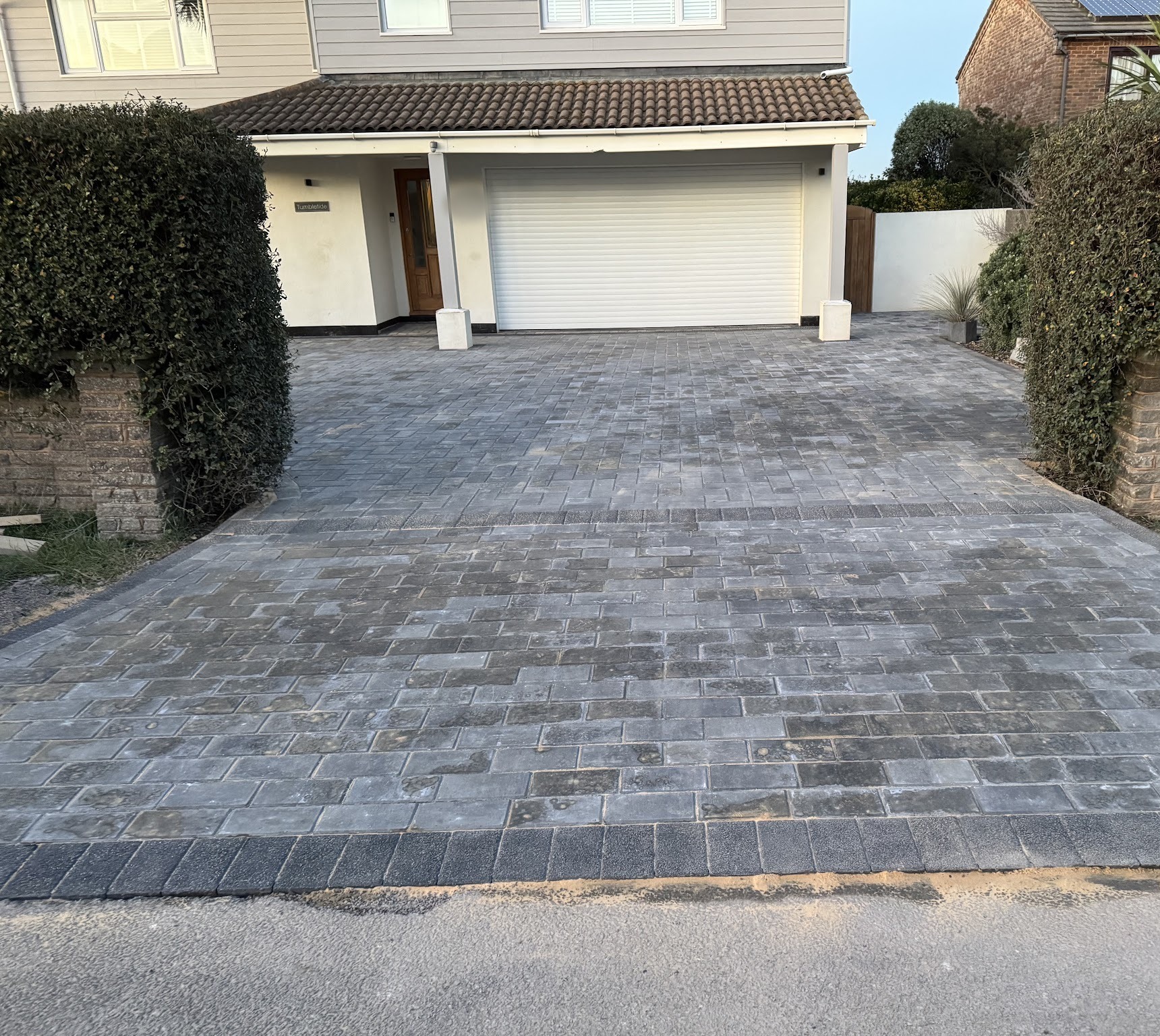 A charcoal block-paved driveway with a darker border in front of a white house